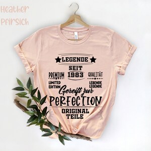 May include: A light pink t-shirt with the text "LEGENDE SEIT 1983" in black. The shirt also features the words "PREMIUM", "QUALIT&Auml;T", "LIMITED EDITION", "Gereift zur PERFECTION", and "ORIGINAL TEILE". The shirt is hanging on a wooden hanger.