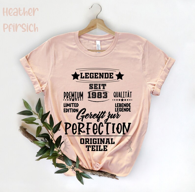 May include: A light pink t-shirt with the text "LEGENDE SEIT 1983" in black. The shirt also features the words "PREMIUM", "QUALIT&Auml;T", "LIMITED EDITION", "Gereift zur PERFECTION", and "ORIGINAL TEILE". The shirt is hanging on a wooden hanger.