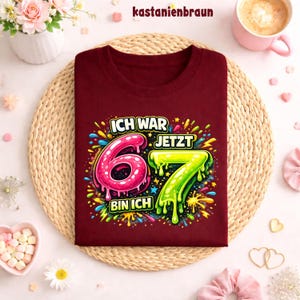 Peut inclure: Un t-shirt bordeaux avec l'inscription "ICH WAR 6 JETZT 7 BIN ICH" dans un design color&eacute; et enfantin. Les chiffres 6 et 7 sont affich&eacute;s en rose et vert. Le t-shirt est pos&eacute; sur un tapis tress&eacute;.