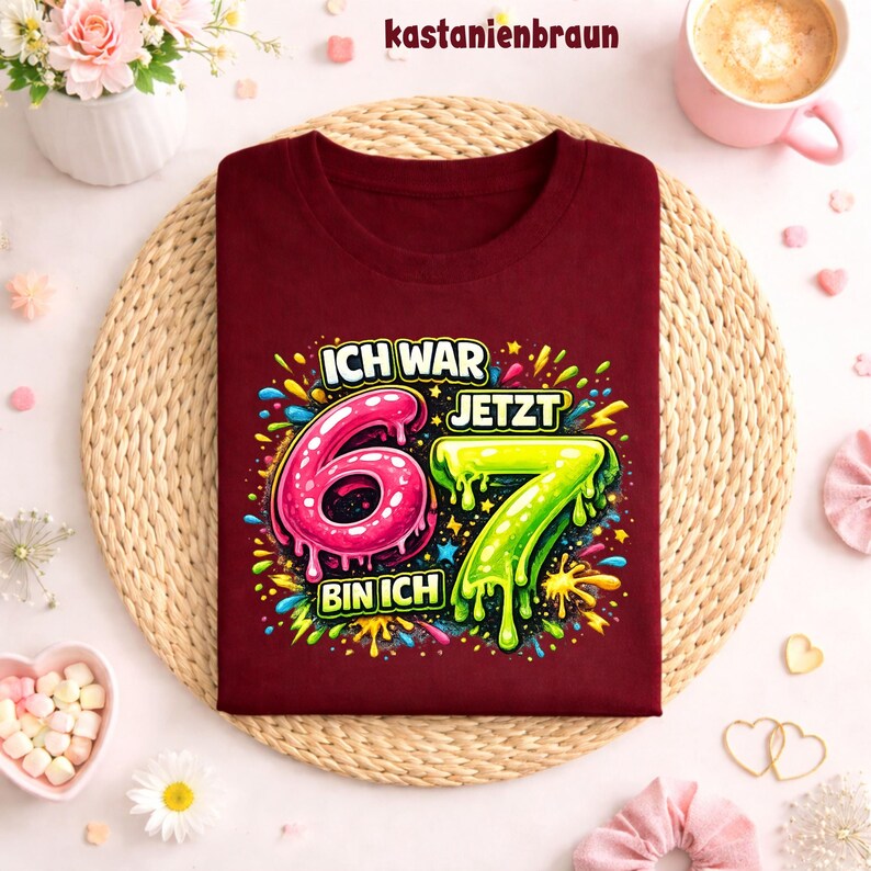 Peut inclure: Un t-shirt bordeaux avec l'inscription "ICH WAR 6 JETZT 7 BIN ICH" dans un design color&eacute; et enfantin. Les chiffres 6 et 7 sont affich&eacute;s en rose et vert. Le t-shirt est pos&eacute; sur un tapis tress&eacute;.
