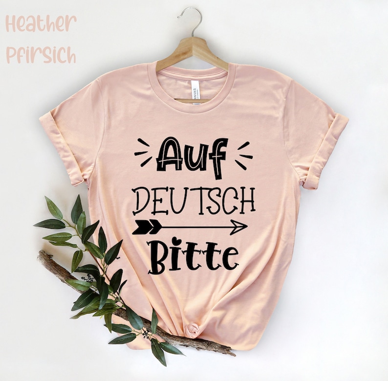 May include: A peach colored t-shirt with black text that reads "Auf Deutsch Bitte" with an arrow pointing to the right.