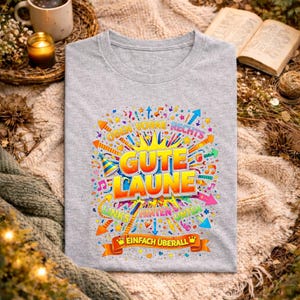 May include: A light gray t-shirt with a colorful graphic that says "Gute Laune" in large letters. The design includes arrows, fireworks, and musical notes. The text "Einfach &Uuml;berall" is at the bottom.