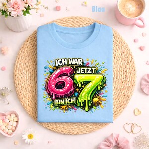 Peut inclure: Un t-shirt bleu clair avec l'inscription "ICH WAR 6 JETZT 7 BIN ICH" dans un design color&eacute; et cartoon. Le chiffre 6 est rose et le chiffre 7 est vert. Le t-shirt est pos&eacute; sur un tapis tress&eacute;.