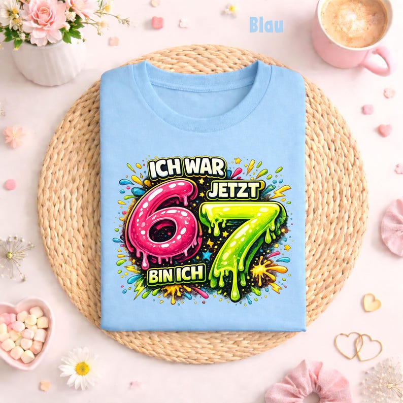 Peut inclure: Un t-shirt bleu clair avec l'inscription "ICH WAR 6 JETZT 7 BIN ICH" dans un design color&eacute; et cartoon. Le chiffre 6 est rose et le chiffre 7 est vert. Le t-shirt est pos&eacute; sur un tapis tress&eacute;.