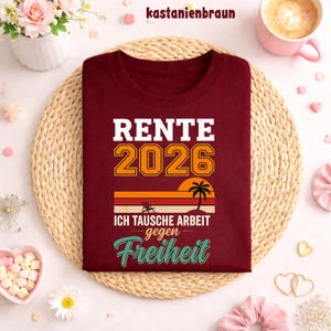 May include: A maroon t-shirt with the text "RENTE 2026" in white and orange, along with the phrase "ICH TAUSCHE ARBEIT gegen Freiheit" and a sunset graphic. The shirt is displayed on a woven mat.