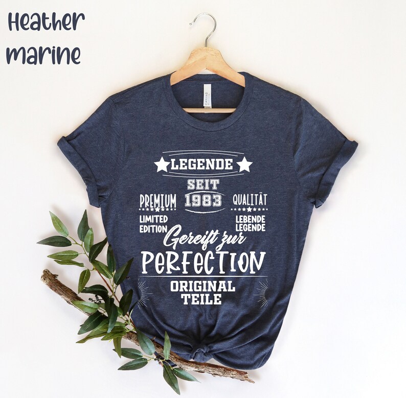 May include: Heather marine navy blue t-shirt with white text. The text includes "LEGENDE SEIT PREMIUM 1983 QUALITAT LIMITED EDITION Gereift zur PERFECTION ORIGINAL TEILE". The shirt is hanging on a wooden hanger.