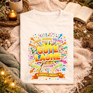 May include: Cream-colored t-shirt with a colorful graphic design. The design includes the words "Gute Laune" in large, yellow and orange letters, surrounded by arrows, stars, and musical notes. The text "Einfach &Uuml;berall" is also present.