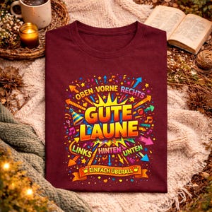 May include: A maroon t-shirt with a colorful graphic design. The design includes the words "Gute Laune" in large yellow letters, surrounded by arrows, musical notes, and other decorative elements. The shirt is displayed on a textured surface.