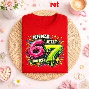 Peut inclure: Un t-shirt rouge avec le texte allemand "ICH WAR 6, JETZT BIN ICH 7" et des graphismes color&eacute;s. Les chiffres 6 et 7 sont affich&eacute;s en rose et vert. Le mot "rot" est &eacute;crit en rouge en haut.