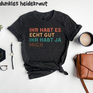 May include: A dark gray t-shirt with the text "Ihr habt es echt gut ihr habt ja mich" in a retro rainbow color scheme.