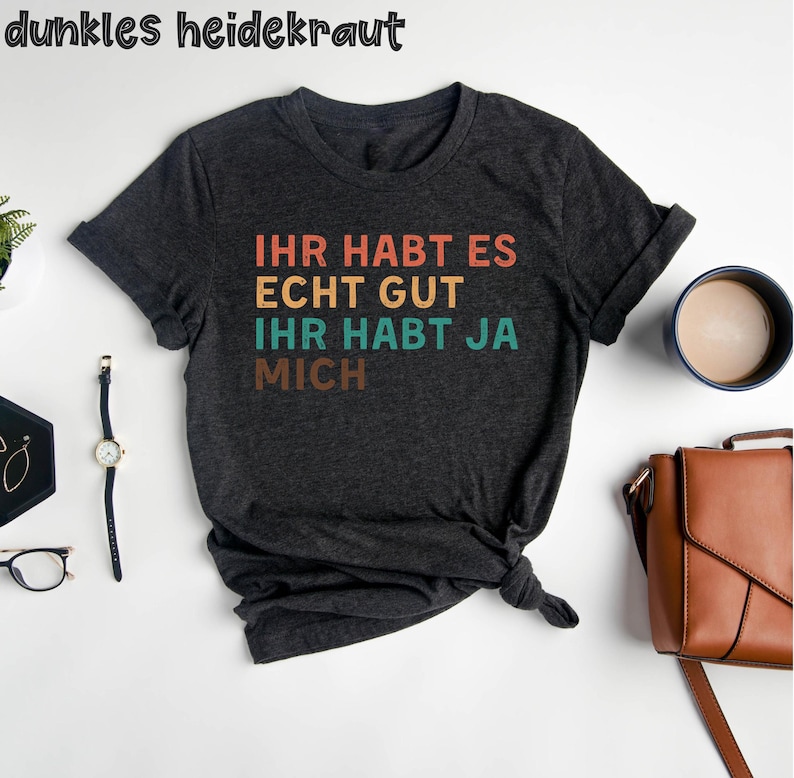 May include: A dark gray t-shirt with the text "Ihr habt es echt gut ihr habt ja mich" in a retro rainbow color scheme.
