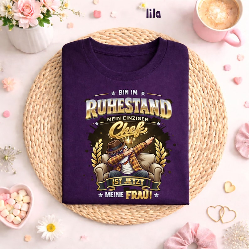 May include: A purple t-shirt with the text "BIN IM RUHESTAND MEIN EINZIGER Chef IST JETZT MEINE FRAU!" in gold and white. The graphic features a cartoon person dabbing while seated. The shirt is displayed on a woven mat.
