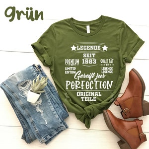 May include: An olive green t-shirt with white text that reads "LEGENDE SEIT PREMIUM 1983 QUALIT&Auml;T LIMITED EDITION Gereift zur PERFECTION ORIGINAL TEILE". The shirt is paired with blue jeans and brown ankle boots.