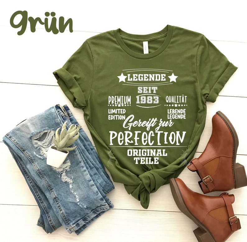 May include: An olive green t-shirt with white text that reads "LEGENDE SEIT PREMIUM 1983 QUALIT&Auml;T LIMITED EDITION Gereift zur PERFECTION ORIGINAL TEILE". The shirt is paired with blue jeans and brown ankle boots.