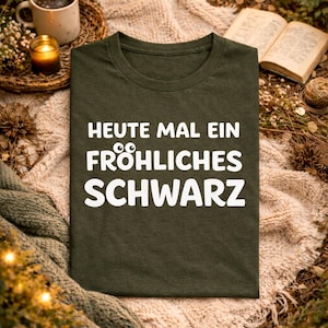 May include: Dark green t-shirt with the German phrase "HEUTE MAL EIN FRÖHLICHES SCHWARZ" in white. The shirt is displayed on a cosy blanket with a cup of coffee, a candle, and an open book in the background.