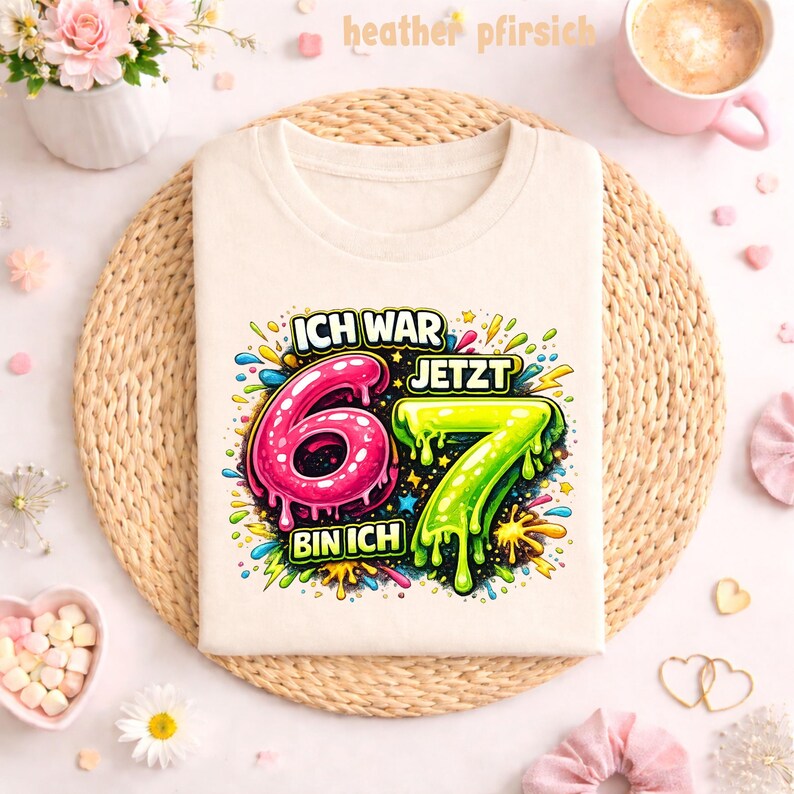 Peut inclure: T-shirt cr&egrave;me avec un graphisme color&eacute; indiquant "ICH WAR 6 JETZT 7 BIN ICH". Les chiffres 6 et 7 sont respectivement roses et verts, avec un motif &eacute;clabouss&eacute;. Le t-shirt est pos&eacute; sur un tapis tiss&eacute;.