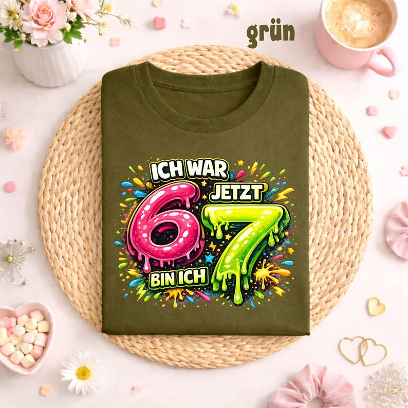 Peut inclure: T-shirt vert olive avec l'inscription "ICH WAR 6 JETZT 7 BIN ICH" dans un design color&eacute;. Le chiffre 6 est rose et le chiffre 7 est vert. Le t-shirt est pos&eacute; sur un tapis tress&eacute;.