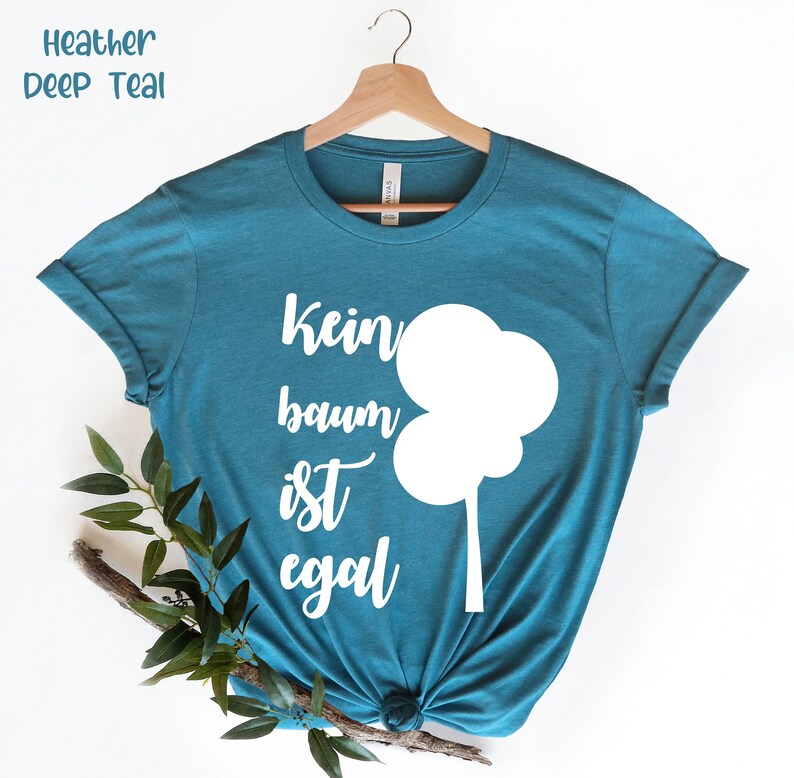 以下が含まれることがあります： 深いティール色のTシャツ。テキスト「Kein baum ist egal」と白い木のグラフィックが特徴です。シャツは白い背景に木製のハンガーに展示されています。「Heather Deep Teal」の文字が左上にあります。