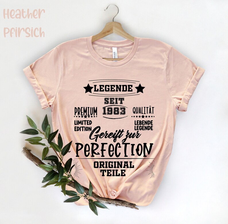 May include: A light pink t-shirt with the text "LEGENDE SEIT 1983 GEREIFT ZUR PERFECTION" in black. The shirt is hanging on a wooden hanger. The text also includes the words "PREMIUM", "QUALIT&Auml;T", "LIMITED EDITION", "LEBENDE LEGENDE", and "ORIGINAL TEILE".
