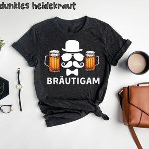 K&ouml;nnte beinhalten: Dunkelgraues T-Shirt mit einer Grafik, die einen wei&szlig;en Hut, eine Sonnenbrille, einen Schnurrbart und eine Fliege zwischen zwei Bierkr&uuml;gen zeigt. Das Wort "BR&Auml;UTIGAM" ist darunter gedruckt. Der Text "dunkles heidekraut" steht oben.