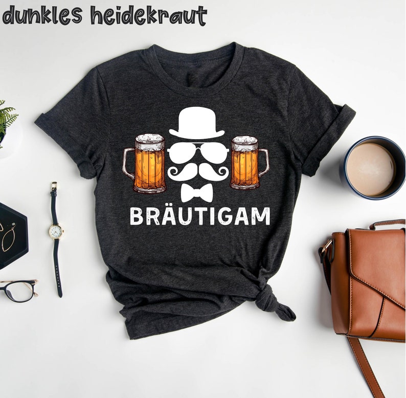 K&ouml;nnte beinhalten: Dunkelgraues T-Shirt mit einer Grafik, die einen wei&szlig;en Hut, eine Sonnenbrille, einen Schnurrbart und eine Fliege zwischen zwei Bierkr&uuml;gen zeigt. Das Wort "BR&Auml;UTIGAM" ist darunter gedruckt. Der Text "dunkles heidekraut" steht oben.