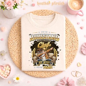 May include: Cream-colored t-shirt with a graphic featuring a man dabbing in a chair, the text reads "BIN IM RUHESTAND MEIN EINZIGER Chef IST JETZT MEINE FRAU!" in gold and black. The shirt is displayed on a woven mat.