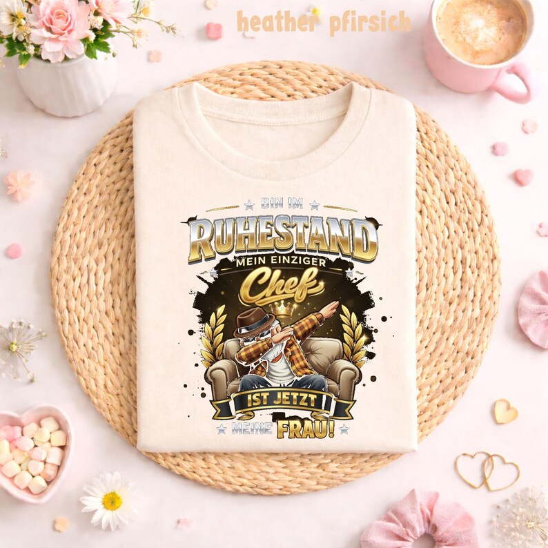 May include: Cream-colored t-shirt with a graphic featuring a man dabbing in a chair, the text reads "BIN IM RUHESTAND MEIN EINZIGER Chef IST JETZT MEINE FRAU!" in gold and black. The shirt is displayed on a woven mat.