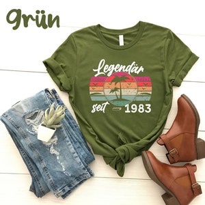 May include: Olive green t-shirt with a retro sunset design featuring palm trees and the text "Legend&auml;r seit 1983". The shirt is paired with blue jeans and brown ankle boots. The word "Gr&uuml;n" is in the top left corner.