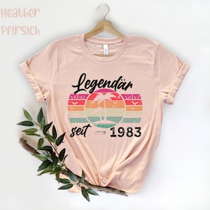 May include: A light pink t-shirt with rolled-up sleeves. The shirt features a retro design with the word "Legend&auml;r" above a colorful sunset graphic with palm trees and the text "seit 1983".