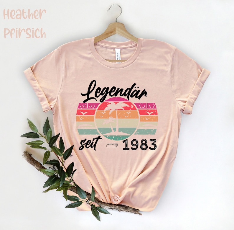 May include: A light pink t-shirt with rolled-up sleeves. The shirt features a retro design with the word "Legend&auml;r" above a colorful sunset graphic with palm trees and the text "seit 1983".
