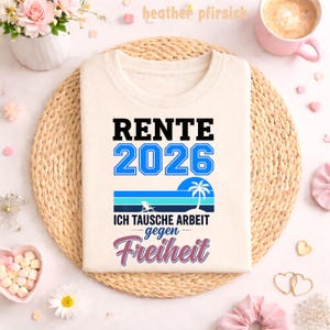 May include: Cream-colored t-shirt with the text "RENTE 2026" in black and blue. Below, a graphic of a beach scene with a palm tree and the words "ICH TAUSCHE ARBEIT gegen Freiheit" in blue and pink. The shirt is displayed on a woven mat.