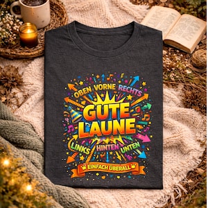 May include: Dark gray t-shirt with a colorful graphic design. The design includes the words "Gute Laune" in large letters, surrounded by arrows, musical notes, and other decorative elements. The text "Oben Vorne Rechts, Links Hinten Unten, Einfach &Uuml;berall" is also included.