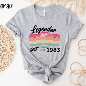 May include: A light gray t-shirt featuring the text "Legend&auml;r seit 1983" above a retro sunset design with a palm tree and birds. The shirt is knotted at the waist.