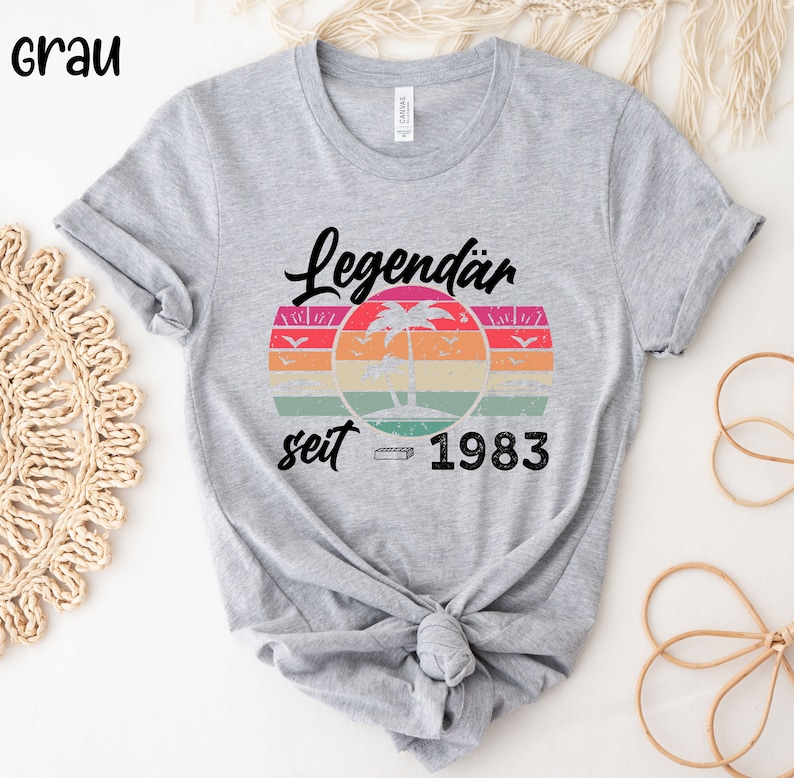 May include: A light gray t-shirt featuring the text "Legend&auml;r seit 1983" above a retro sunset design with a palm tree and birds. The shirt is knotted at the waist.