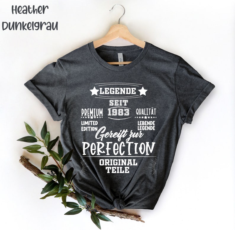 May include: Heather gray t-shirt with white text. The text includes "LEGENDE SEIT 1983", "Gereift zur PERFECTION", and "ORIGINAL TEILE". The shirt is hanging on a wooden hanger.