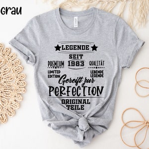 May include: A gray t-shirt with black text. The text includes "LEGENDE SEIT 1983", "Gereift zur PERFECTION", and "ORIGINAL TEILE". The shirt is knotted at the bottom.
