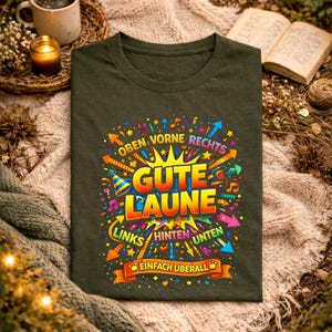 May include: Dark green t-shirt with a colorful graphic design. The design includes the words "Gute Laune" in large, yellow and orange letters, surrounded by arrows, musical notes, and other decorative elements. The text "Oben Vorne Rechts" and "Links Hinten Unten" are also included.