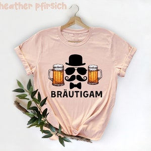 K&ouml;nnte beinhalten: Hellrosa T-Shirt mit einem Grafikdesign. Das Design zeigt einen schwarzen Bowlerhut, eine Sonnenbrille, einen Schnurrbart, eine Fliege und zwei Bierkr&uuml;ge. Das Wort "BR&Auml;UTIGAM" ist darunter gedruckt.