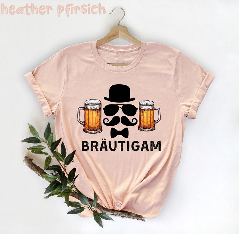 K&ouml;nnte beinhalten: Hellrosa T-Shirt mit einem Grafikdesign. Das Design zeigt einen schwarzen Bowlerhut, eine Sonnenbrille, einen Schnurrbart, eine Fliege und zwei Bierkr&uuml;ge. Das Wort "BR&Auml;UTIGAM" ist darunter gedruckt.
