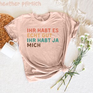 May include: A light pink t-shirt with the text "Ihr habt es echt gut ihr habt ja mich" in a rainbow color scheme.