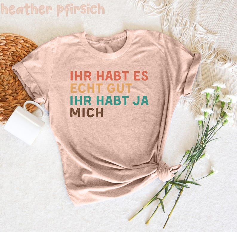 May include: A light pink t-shirt with the text "Ihr habt es echt gut ihr habt ja mich" in a rainbow color scheme.