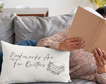 May include: White throw pillow with the text "Bookmarks Are For Quitters" in black script font. A stack of books with a floral design is pictured next to the text.
