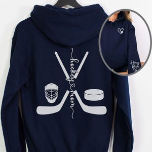 K&ouml;nnte beinhalten: Marineblauer Hoodie mit einem wei&szlig;en Hockeyschl&auml;ger- und Puck-Design auf der R&uuml;ckseite. Der Text "hockey & mom" ist vertikal auf der R&uuml;ckseite des Hoodies geschrieben.