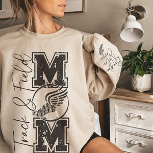 May include: Beige sweatshirt with the words "Track & Field Mom" in black, with a distressed design. The sweatshirt features the word "MOM" in large block letters and a track and field emblem. The sleeve has the names "Braden, Nolan, Carson".