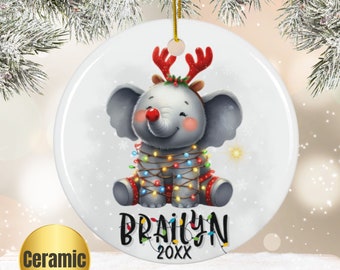 Może przedstawiać: Biała ceramiczna ozdoba choinkowa przedstawiająca kreskówkowego słonia w czerwono-zielonym opaską na głowę z rogami renifera i owiniętego kolorowymi światełkami choinkowymi. Słoń siedzi i uśmiecha się. Ozdoba jest spersonalizowana imieniem "Brailyn" i rokiem "20XX".