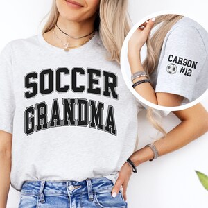 May include: Gray t-shirt with the text "SOCCER GRANDMA" in black. The sleeve of the shirt has a white soccer ball with the text "CARSON #12" in black.