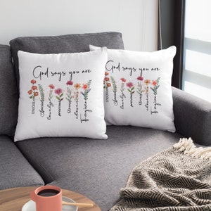 Pode incluir: Duas almofadas brancas com desenhos florais e o texto "Deus diz que voc&ecirc; &eacute;" com uma lista de atributos positivos abaixo. As almofadas est&atilde;o em um sof&aacute; cinza.