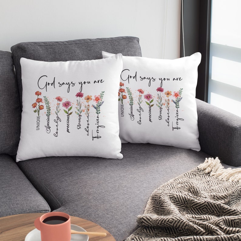 Pode incluir: Duas almofadas brancas com desenhos florais e o texto "Deus diz que voc&ecirc; &eacute;" com uma lista de atributos positivos abaixo. As almofadas est&atilde;o em um sof&aacute; cinza.