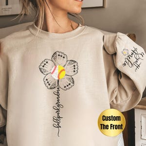 May include: A beige sweatshirt with a black and white graphic of a softball glove flower with a yellow softball in the center. The text "ballpark grandma" is written vertically below the flower. The text "Custom The Front" is written in a gold box on the bottom right corner of the sweatshirt.