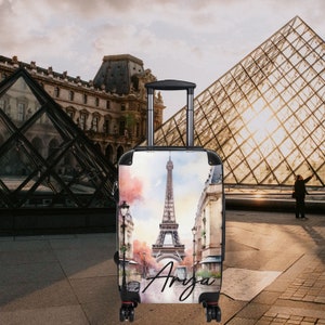 May include: Black carry-on suitcase with a watercolor design of the Eiffel Tower in Paris, France. The suitcase has a black handle and wheels. The text "Anya" is written on the front of the suitcase.